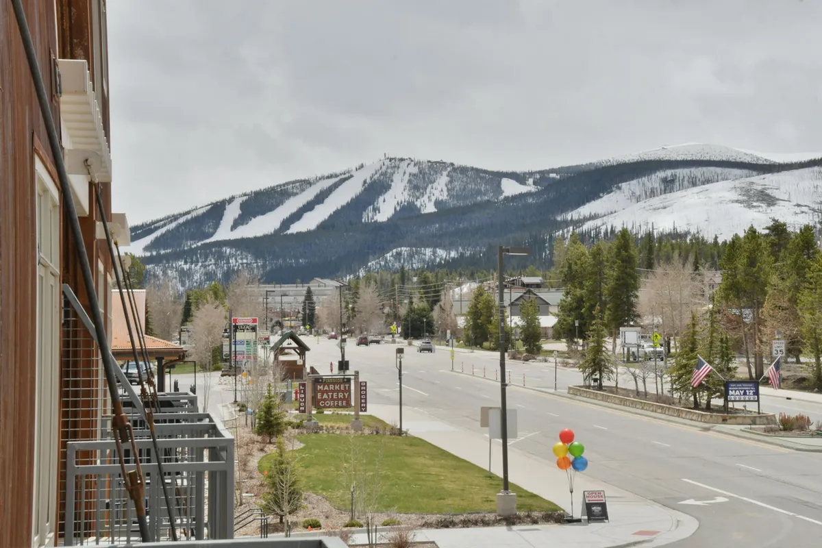 A picturesque mountain town view featuring snow-capped peaks, signage, colorful balloons, and a tree-lined street. The scene evokes a sense of adventure and welcoming community, with visual cues for both winter and summer activities.