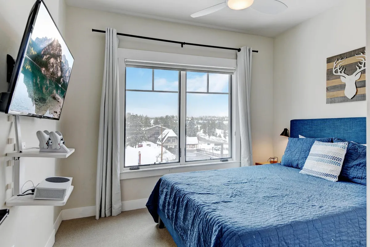A serene bedroom with a bed featuring a plush blue quilt is oriented toward a large window, offering views of a snowy landscape. A wall-mounted TV is visible, providing both entertainment and modern comforts. The atmosphere is calm, inviting restful relaxation after a day in the mountains.