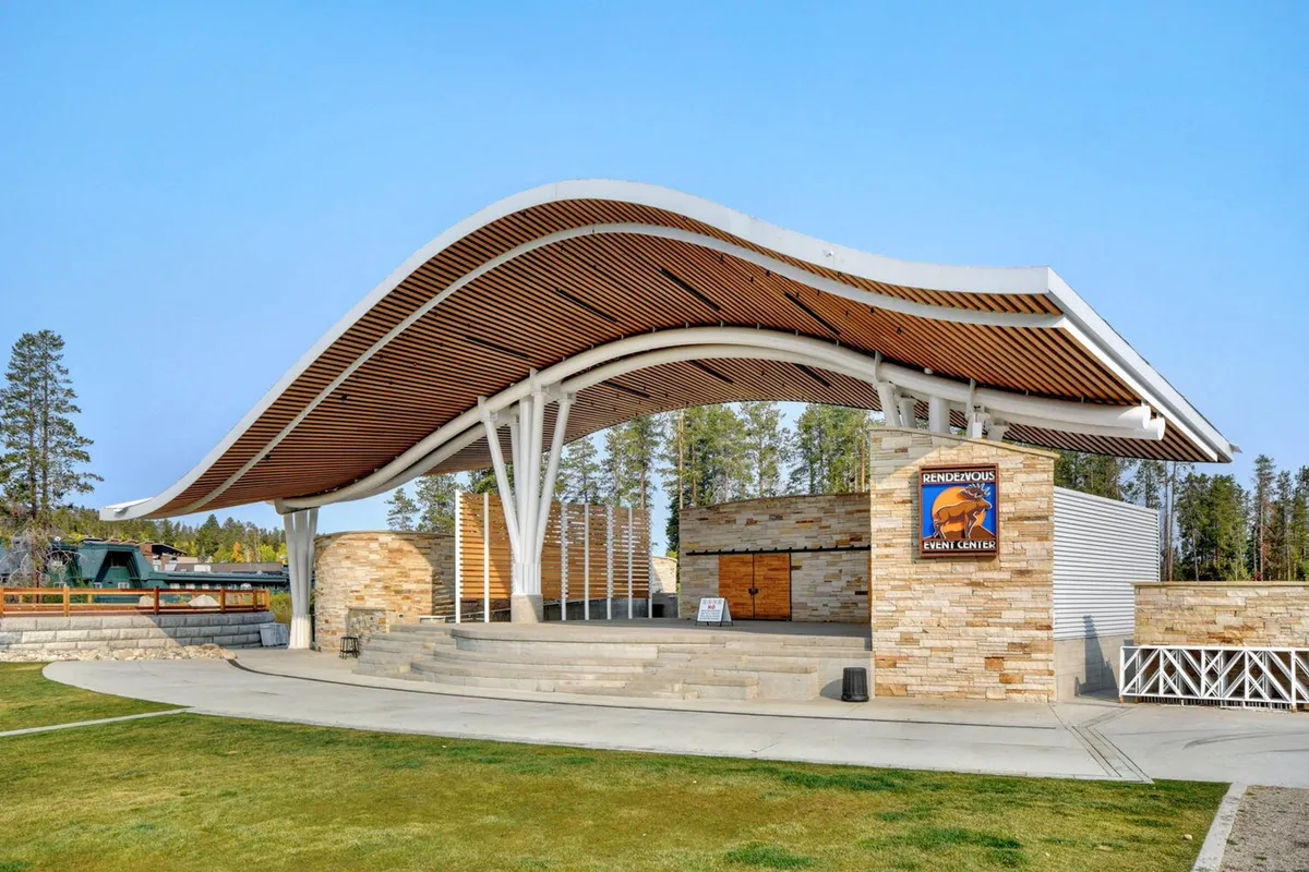 The distinctive curved wooden roof structure of Hideaway Park amphitheater, with a stone façade and open stage area. People gather for a lively outdoor event, reflecting the energy of community gatherings, concerts, and festivals.