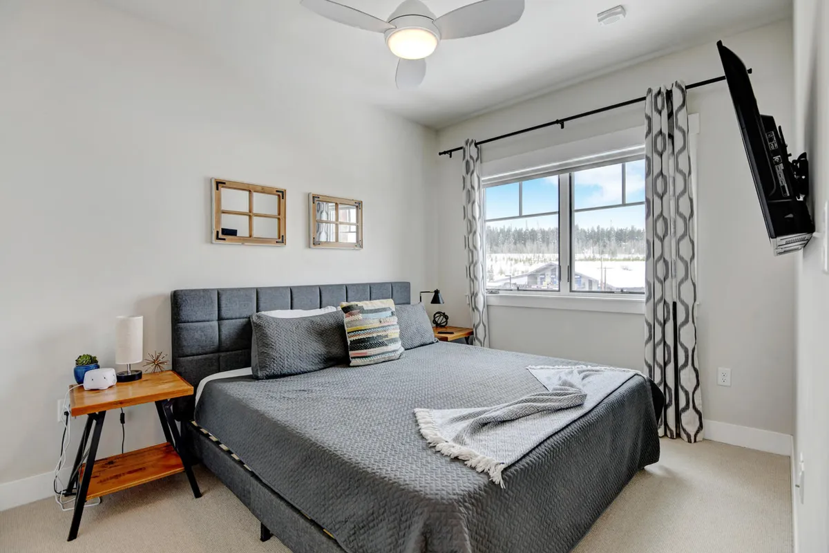 A comfortable bedroom with a large bed, window letting in natural light, decorative mirrors, a nightstand, and a ceiling fan. The mood is cozy and serene, ideal for restful nights or quiet moments during the day.