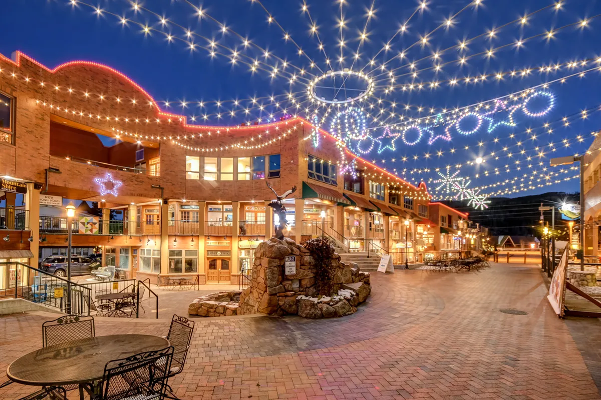 Set under string lights and decorative stars, this image features a stone fountain, outdoor seating, and people enjoying al fresco dining and nighttime strolls. The space feels joyful and active, suggesting a community hub that hosts festivals, markets, and evening gatherings.