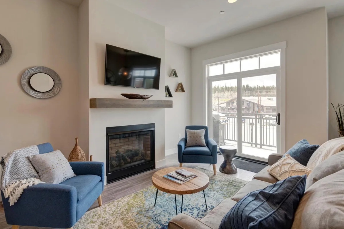 A cozy living area centered around a gas fireplace, with a comfortable sofa, chairs, coffee table, and a wall-mounted television. Large windows provide soft natural light, and the ambiance is both welcoming and intimate.