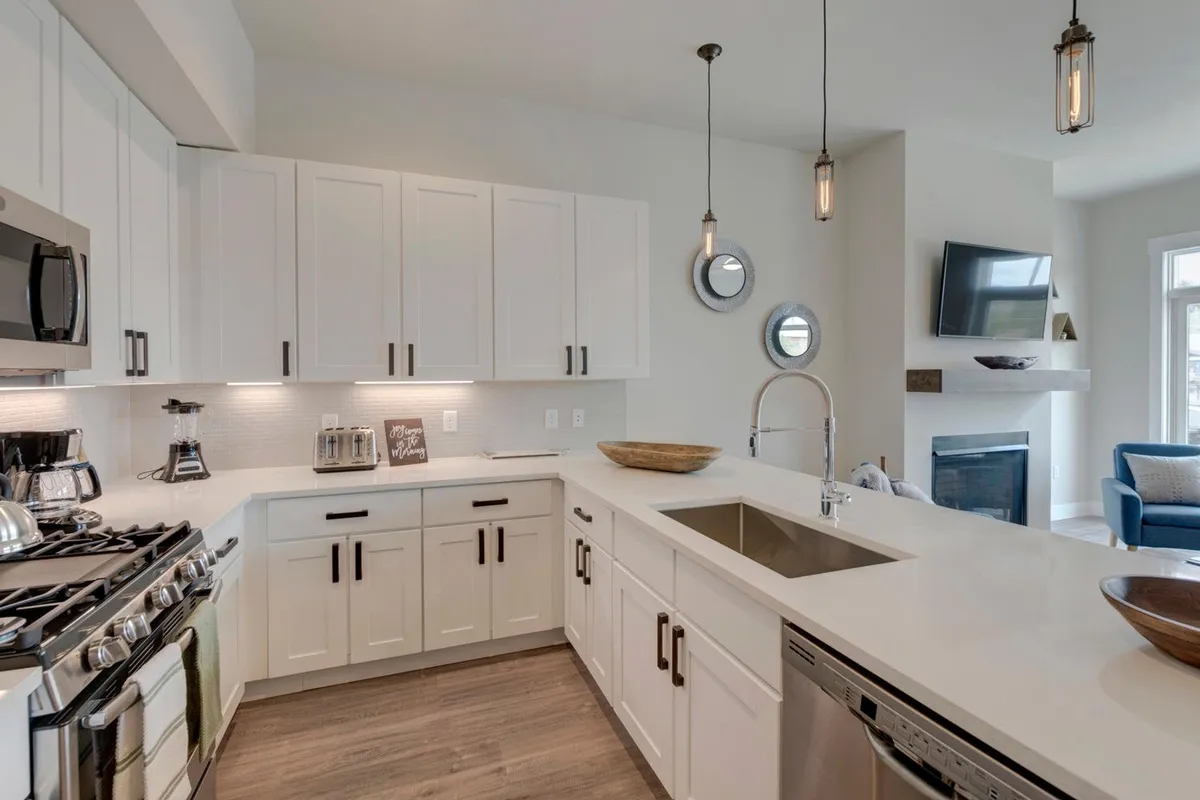 A modern kitchen with stainless steel appliances, crisp white cabinetry, an inviting kitchen island with pendant lighting, and a glimpse of a cozy fireplace in the backdrop. The setup is both functional and stylish, ideal for cooking and entertaining.