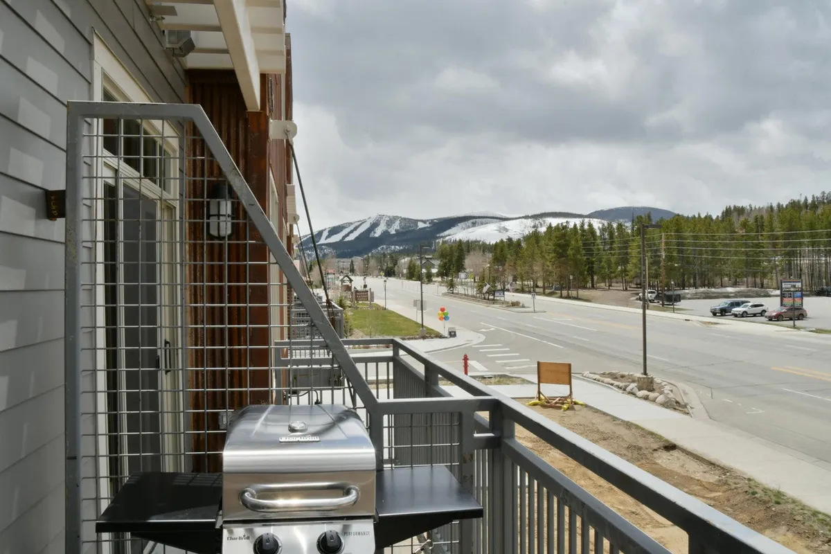 This exterior image features a private balcony area with a barbecue grill, scenic mountain views, and a roadway below. The setting evokes a casual, relaxed atmosphere—perfect for morning coffee or evening grilling while taking in the Rockies.