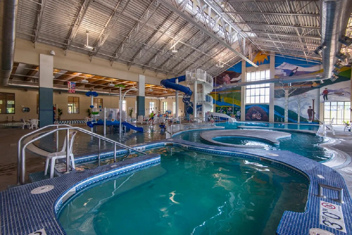 A colorful and energetic indoor aquatic center with water slides, play areas, and a spacious pool. The space is filled with children and families enjoying swimming, splashing, and relaxing by the pool, with a lively mural on the wall adding to the cheerful atmosphere.