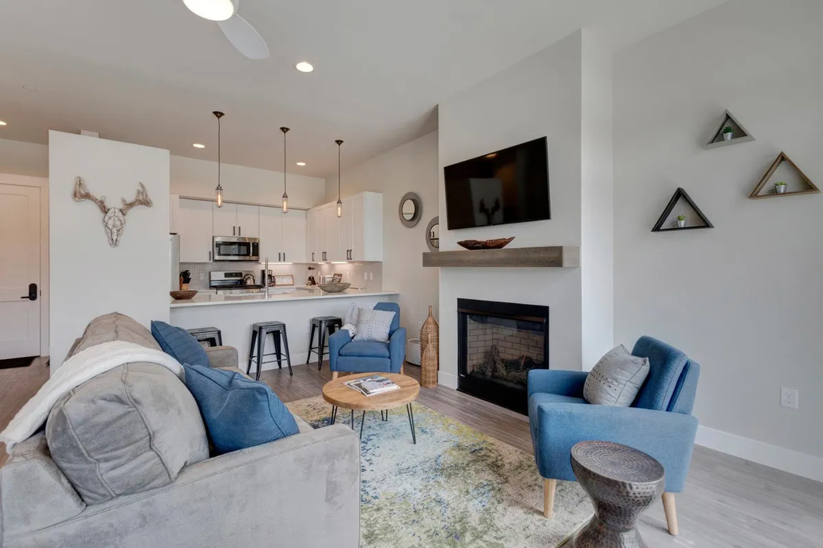 The image showcases a stylish open-concept living area featuring a plush gray sofa, vibrant blue accent chairs, a cozy fireplace, and a modern kitchen in the background. The room exudes warmth and comfort, perfect for gathering with loved ones after a day of Winter Park adventures.