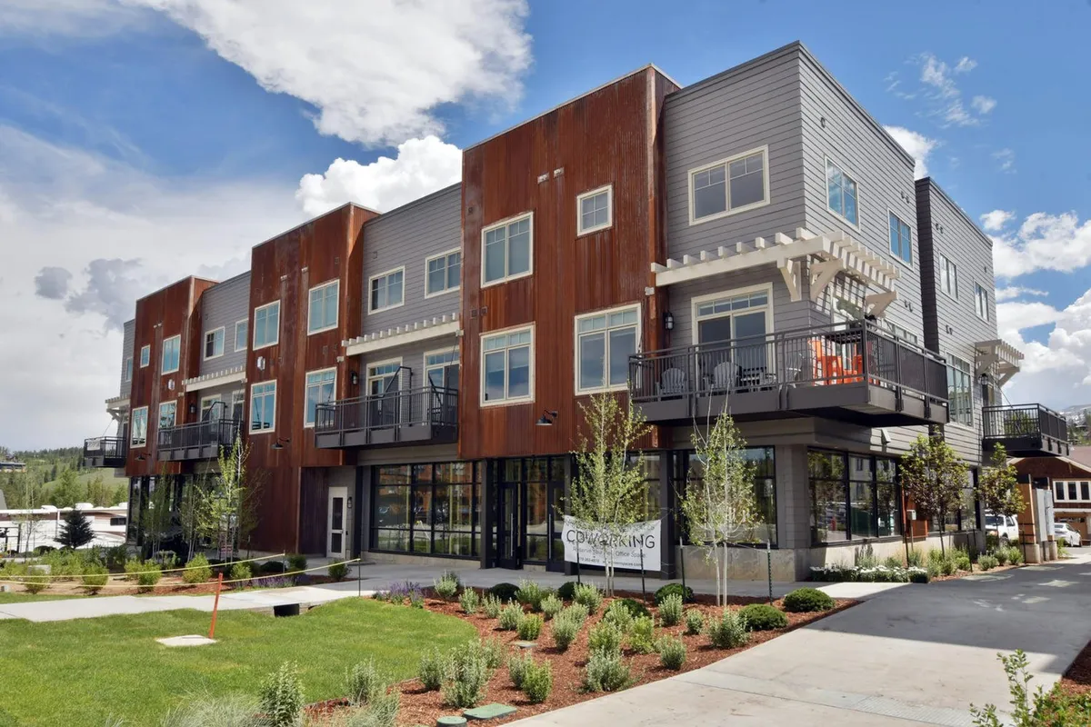 A modern multi-story residential building with stylish architecture, green landscaping, spacious balconies, and large windows. Soft evening light bathes the facade, and people can be seen enjoying the balconies and the communal spaces below.