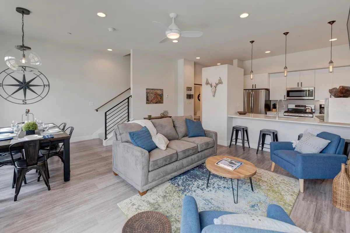 An inviting open-plan space featuring a plush sofa, a large dining table perfect for family gatherings, and a well-equipped kitchen island set against modern decorative touches. Natural light streams in, and the zone is clearly designed for both relaxation and entertaining.